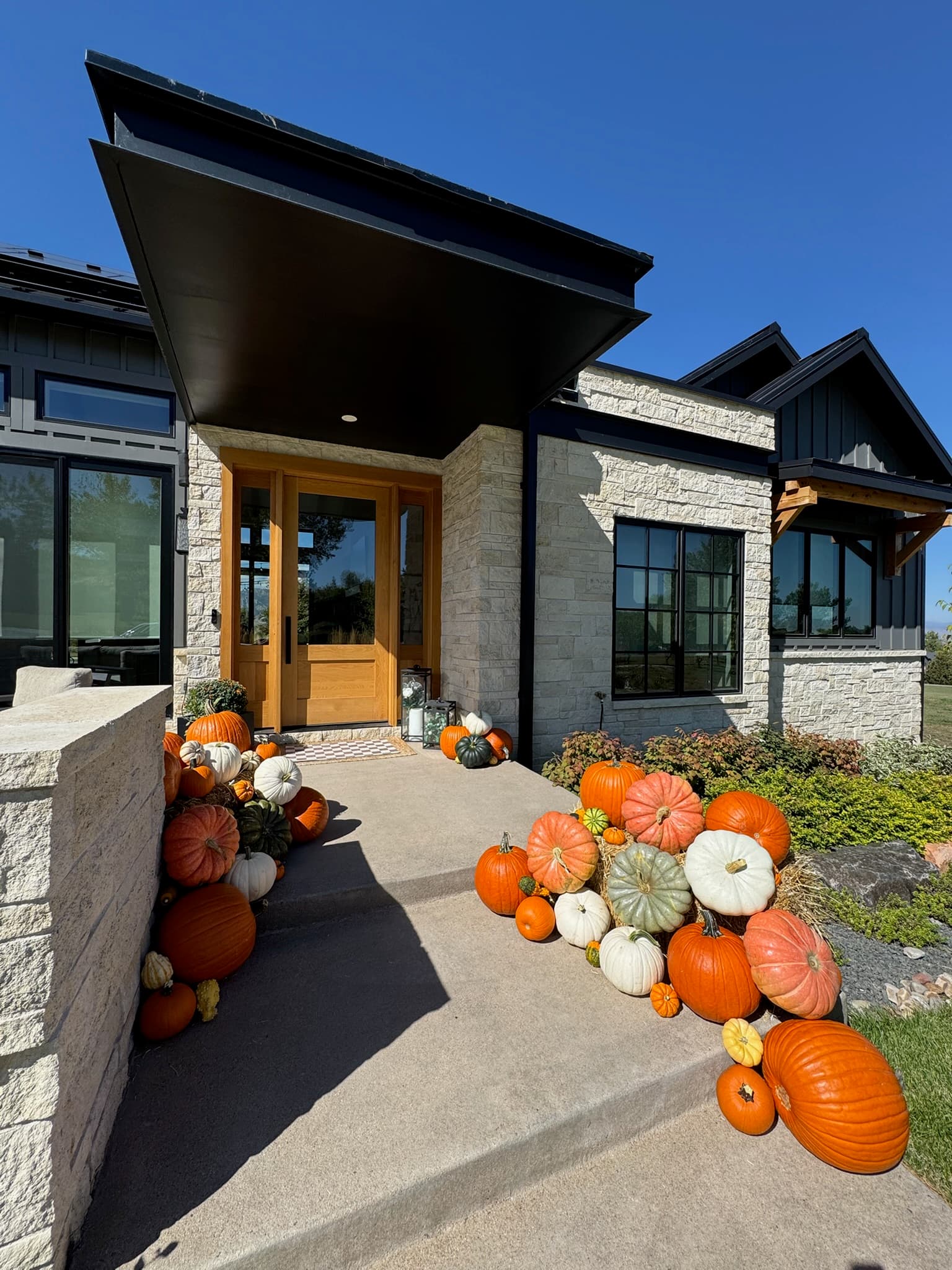 Modern stacked-stone entry with a light wood door and pumpkin clusters on both sides of the walkway