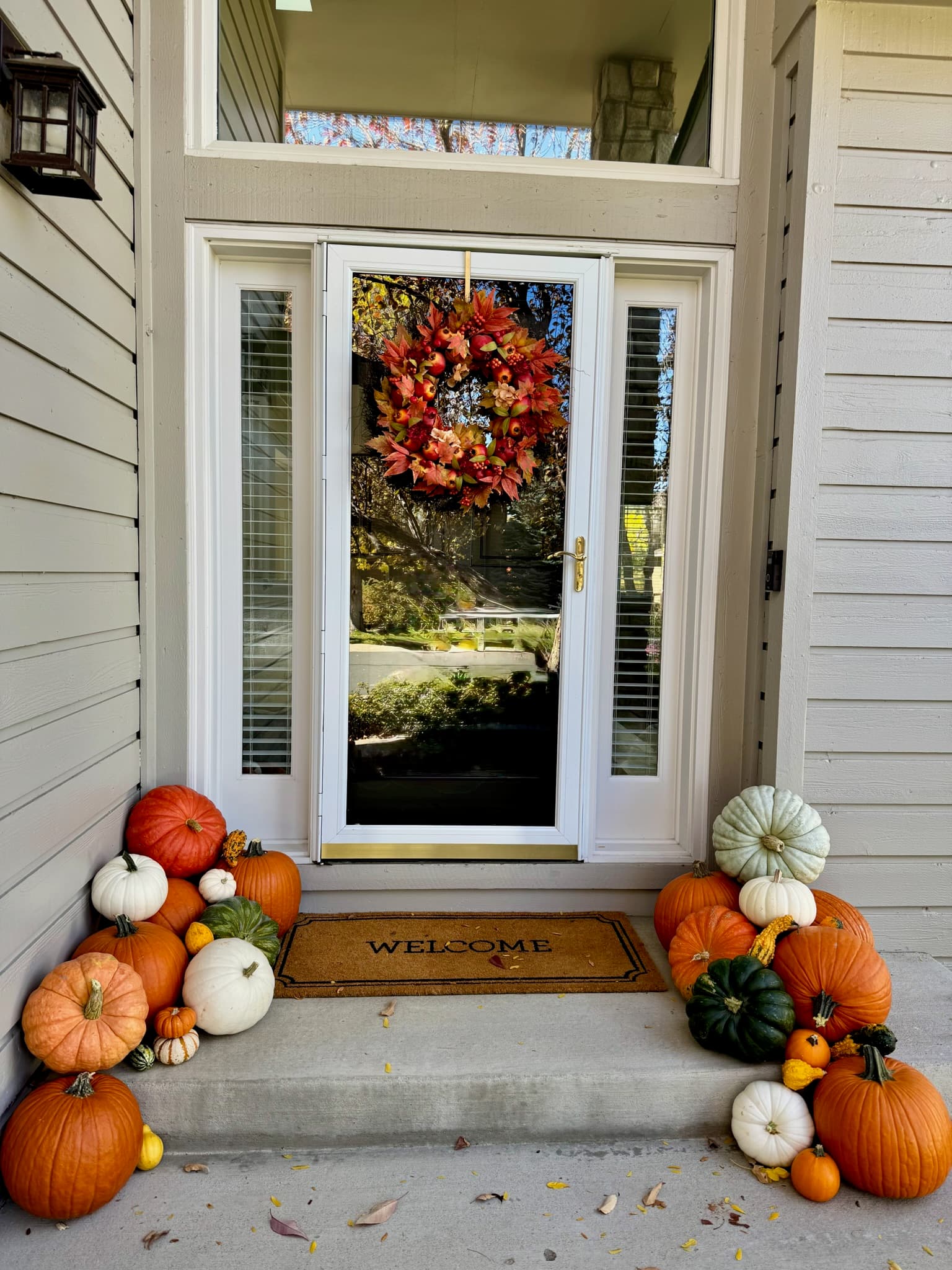 A beige house with a white storm door, a fall leaf wreath, and pumpkin clusters on both sides of a WELCOME mat