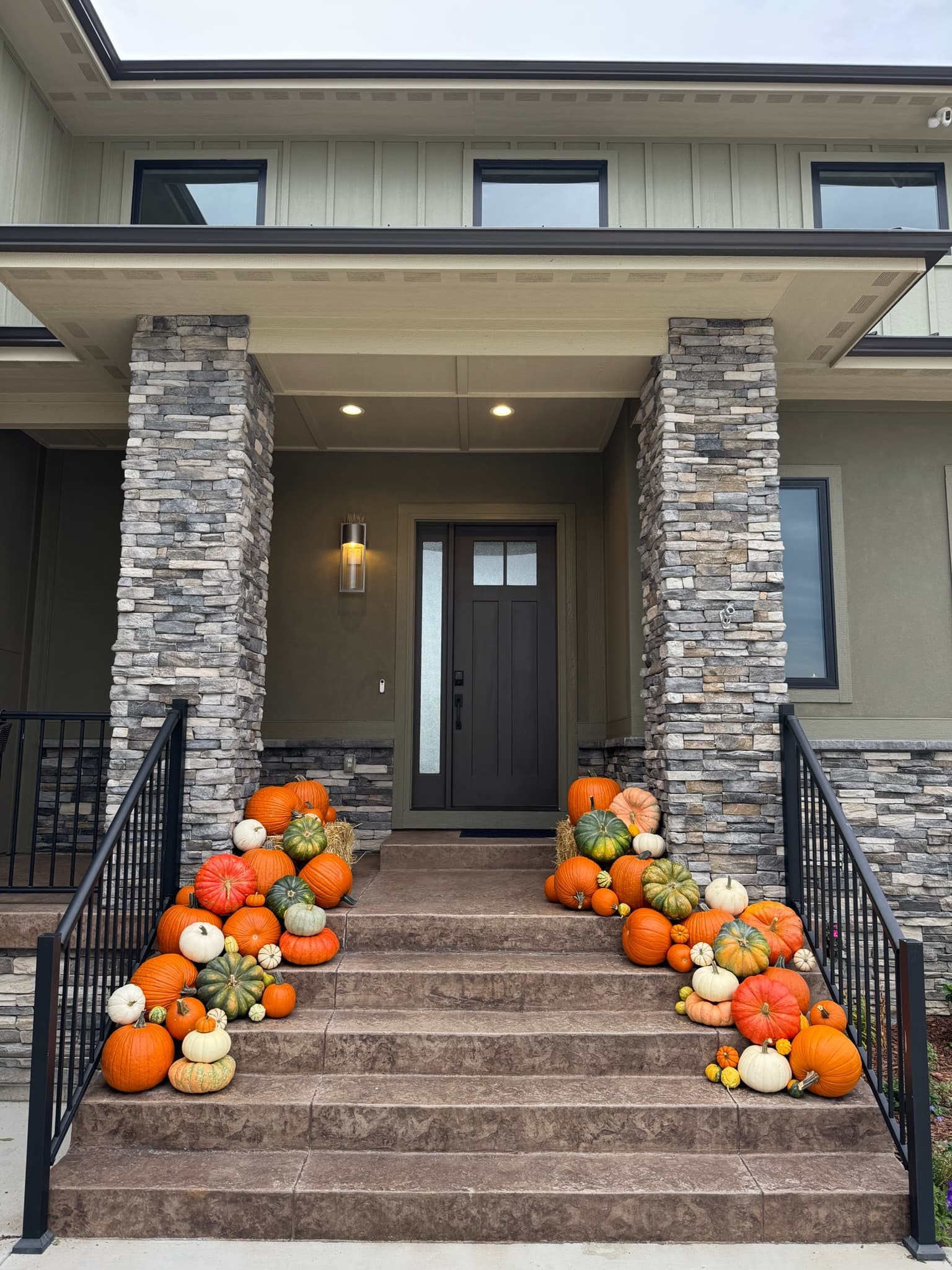 A modern porch with stacked-stone pillars and a dark brown door, with pumpkins cascading down both sides of the steps from hay bales at the top